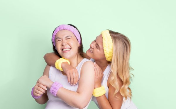 Two girls laughing wearing purple and yellow skincare headbands and matching spa wristbands