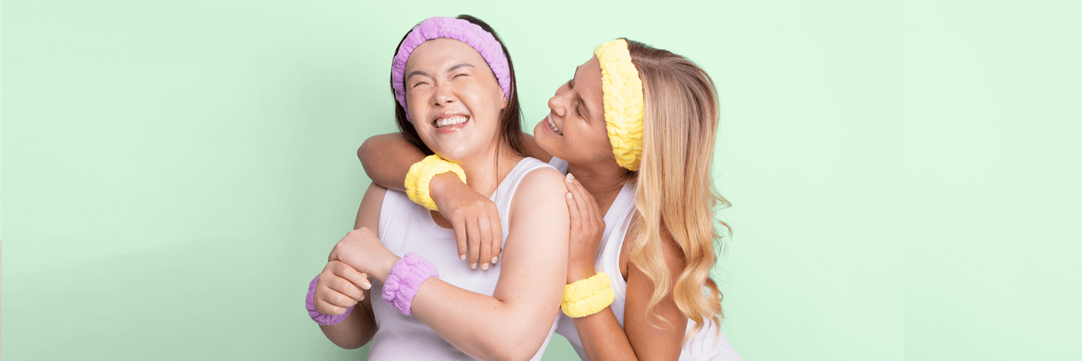 Two girls laughing wearing purple and yellow skincare headbands and matching face washing wrist towels